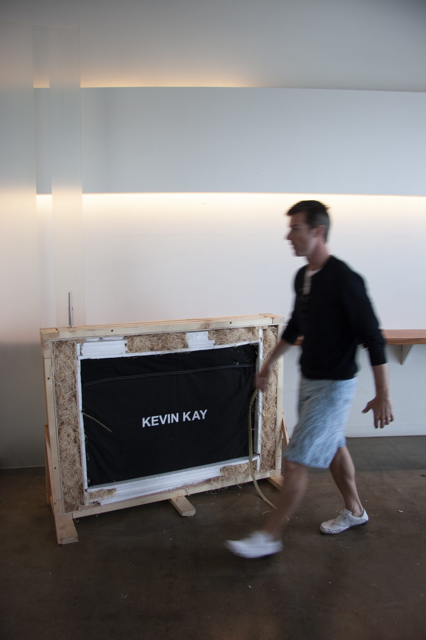 Fine art photographer Kevin Kay walking past a crated artwork in a gallery, the crate front printed with his name.